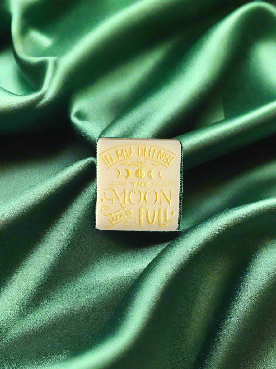 A wax melt with the text 'In my defense, the MOON WAS FULL' on it, placed on a green textured surface.
