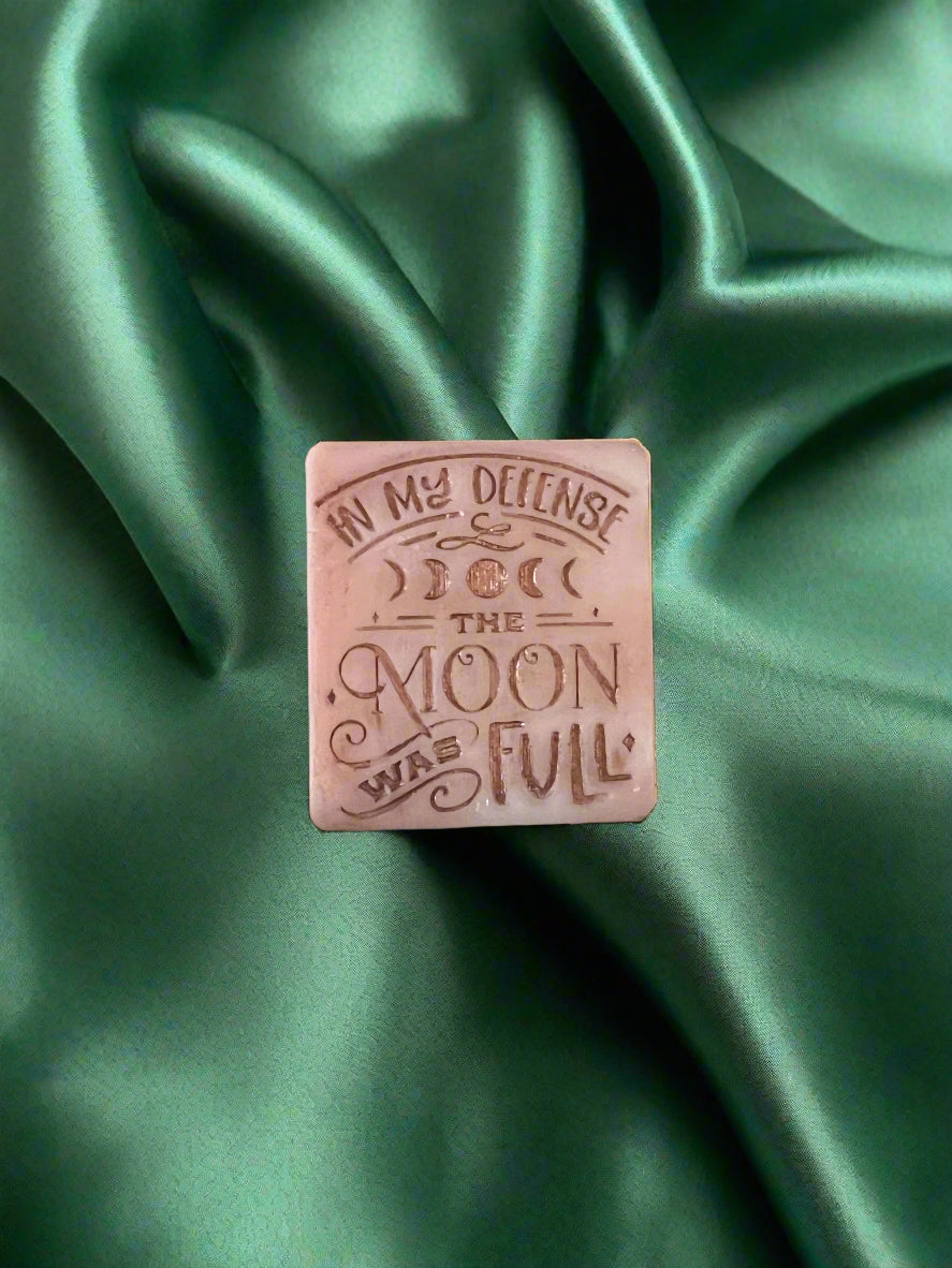 A wax melt with the text 'In my defense, the MOON WAS FULL' on it, placed on a green textured surface.