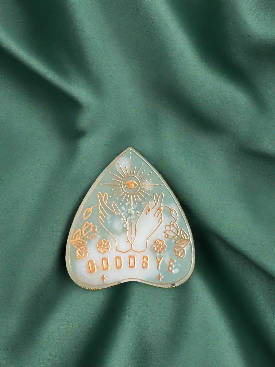 A wax melt in the shape of a planchette with a floral pattern and the word 'GOODBYE' printed on it, placed on a green textured surface.