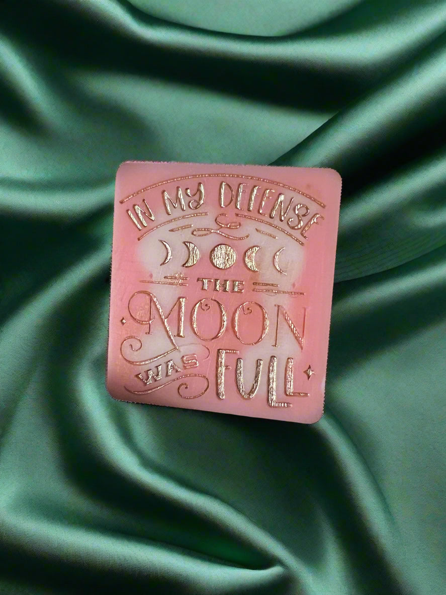 A wax melt with the text 'In my defense, the MOON WAS FULL' on it, placed on a green textured surface.