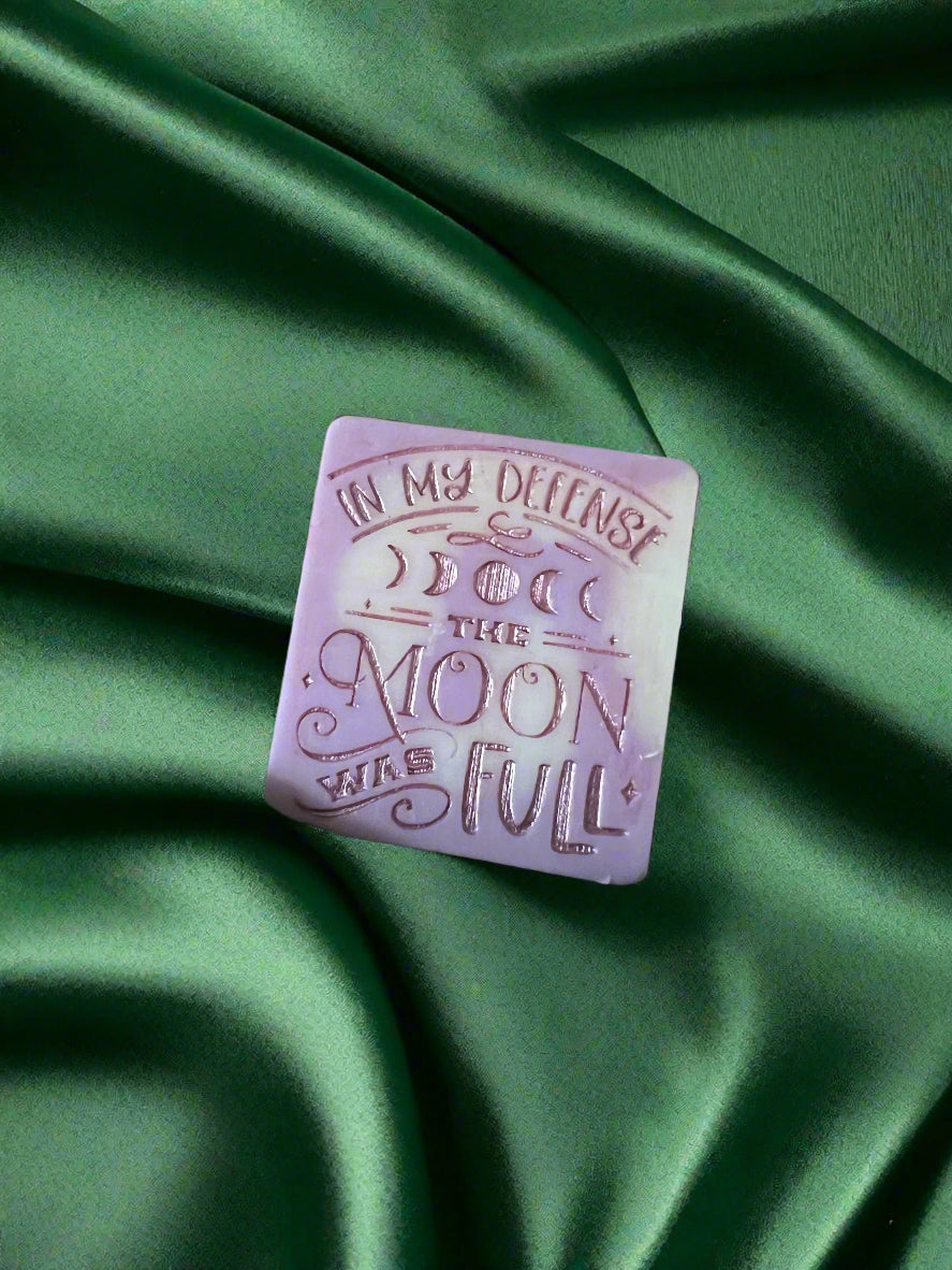 A wax melt with the text 'In my defense, the MOON WAS FULL' on it, placed on a green textured surface.
