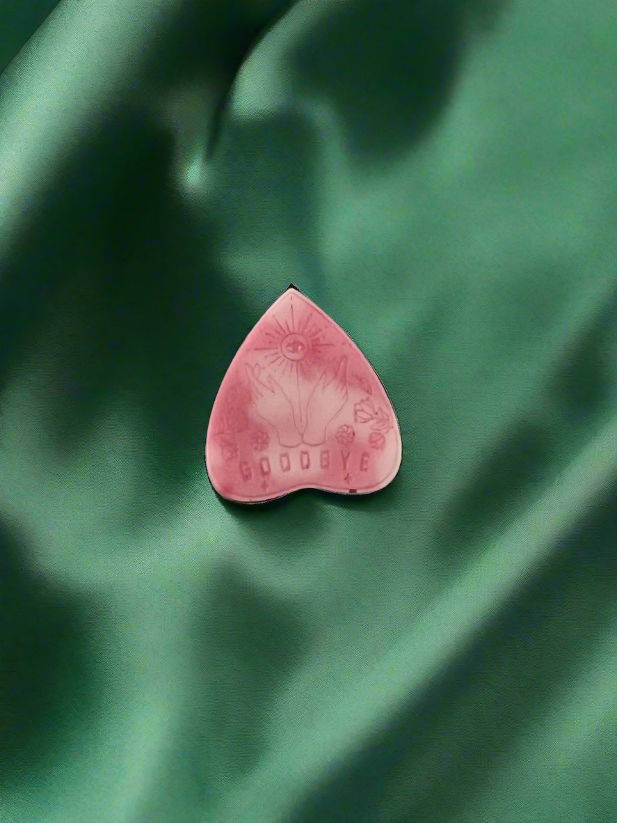 A wax melt in the shape of a planchette with a floral pattern and the word 'GOODBYE' printed on it, placed on a green textured surface.