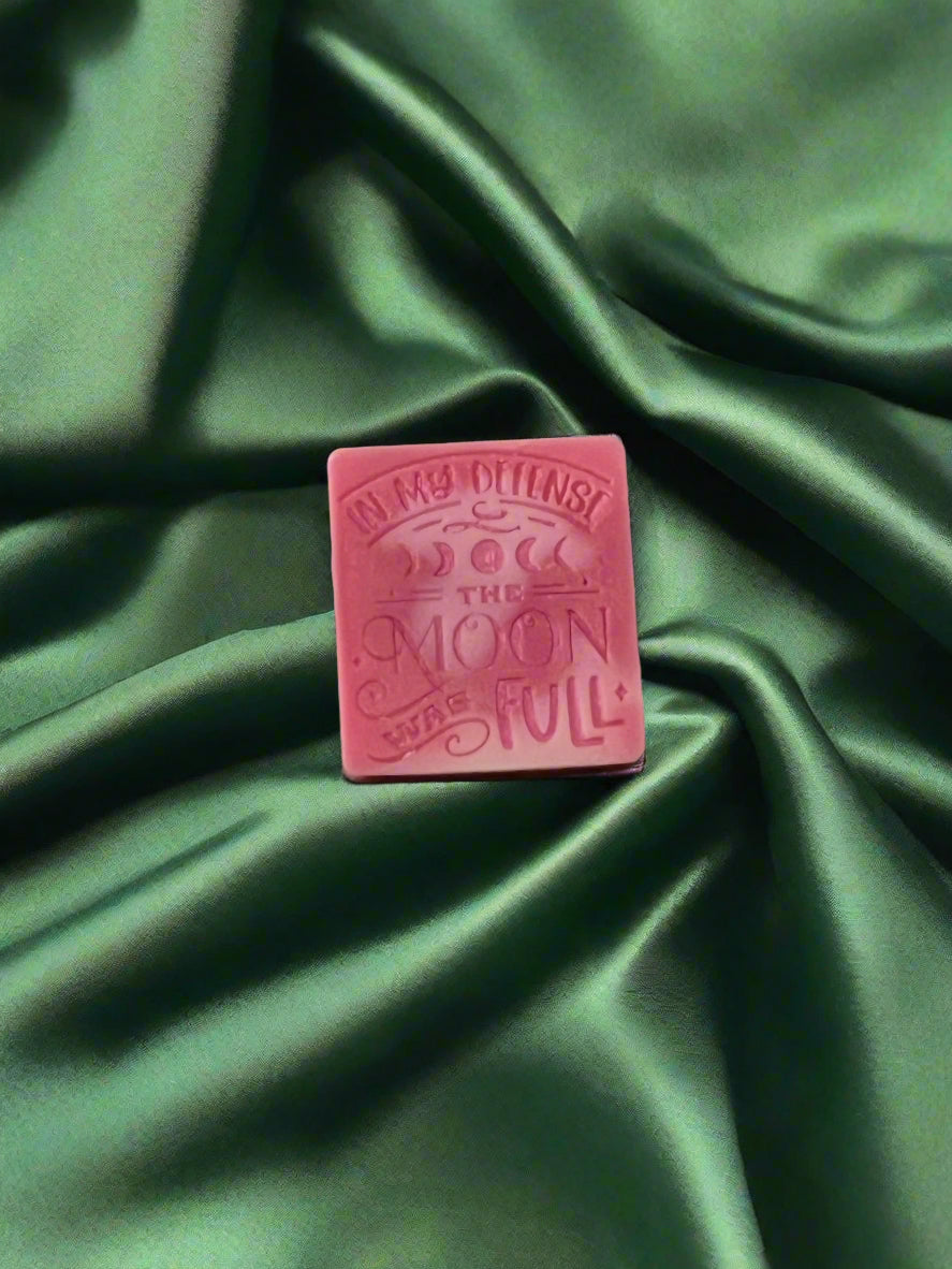 A wax melt with the text 'In my defense, the MOON WAS FULL' on it, placed on a green textured surface.