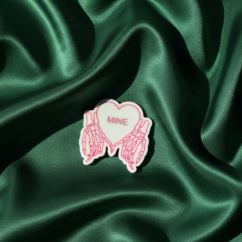 Heart-shaped pin with 'MINE' text on a green satin background