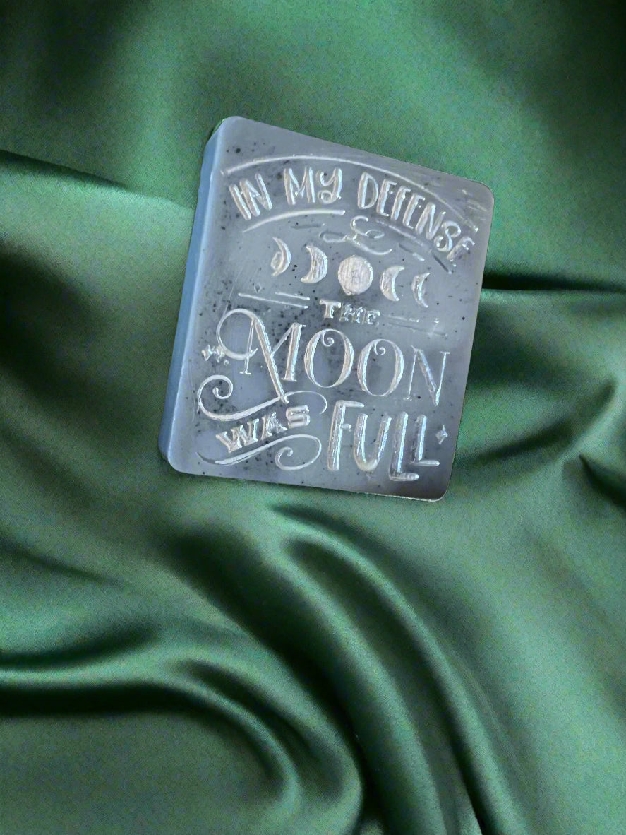 A wax melt with the text 'In my defense, the MOON WAS FULL' on it, placed on a green textured surface.