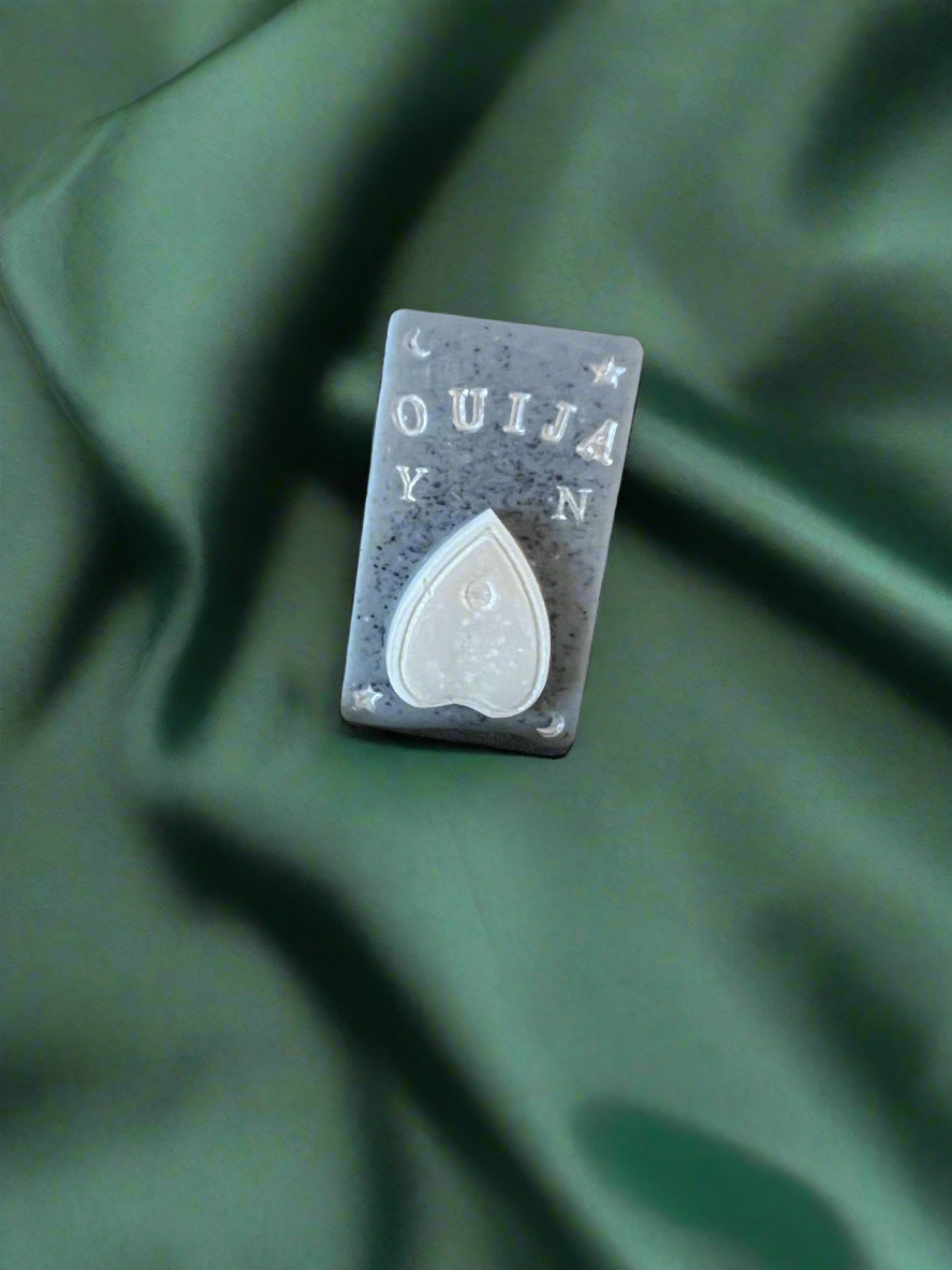 A white wax melt in the shape of a planchette with the word 'OUIJA' above it, placed on a dark green fabric surface.