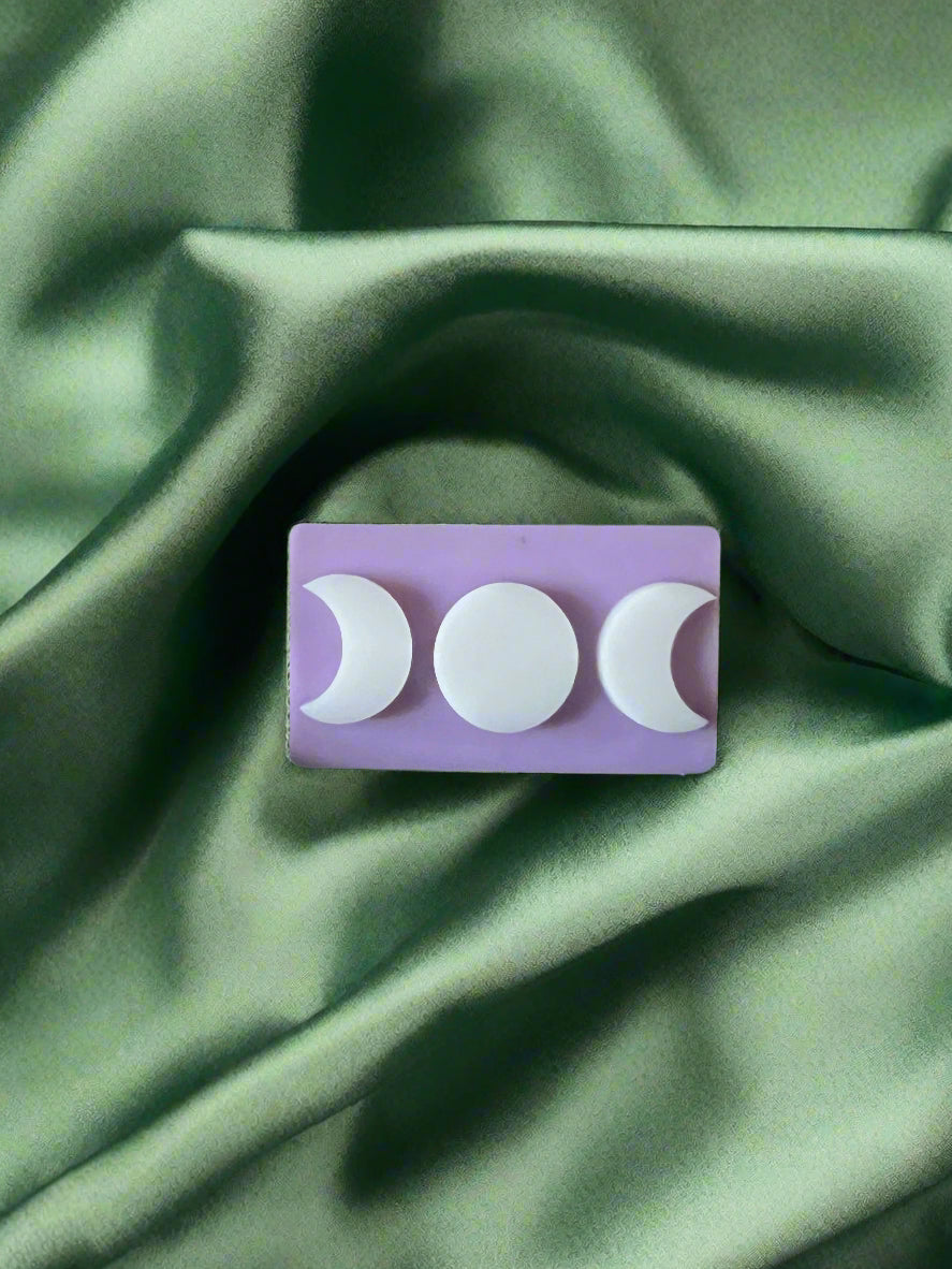 A white wax melt with a triple moon design on a green textured surface.