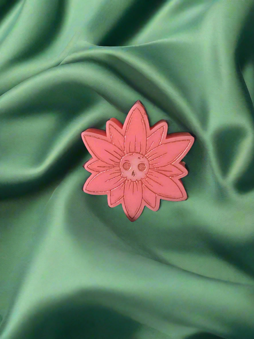 A white wax melt in the shape of a flower with a skull design at the center, placed on a green fabric surface.