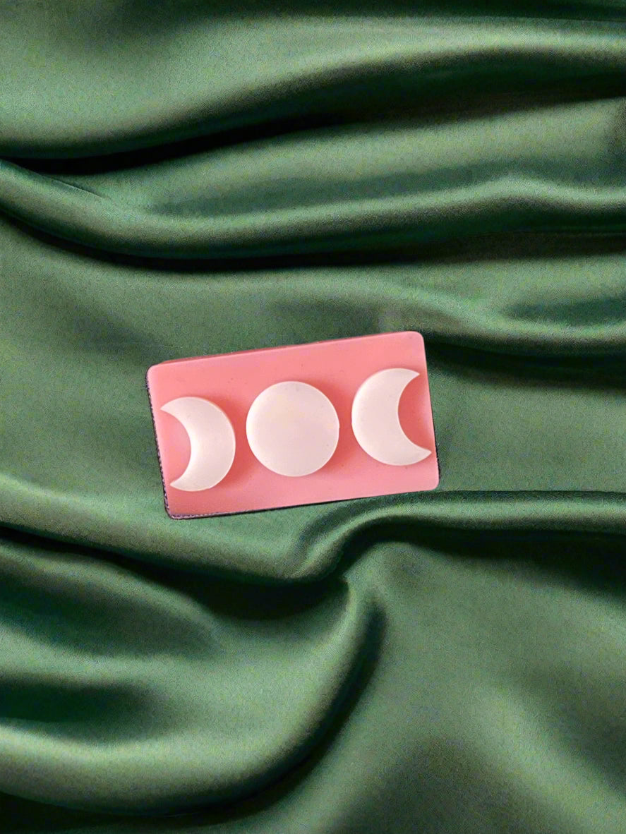 A white wax melt with a triple moon design on a green textured surface.