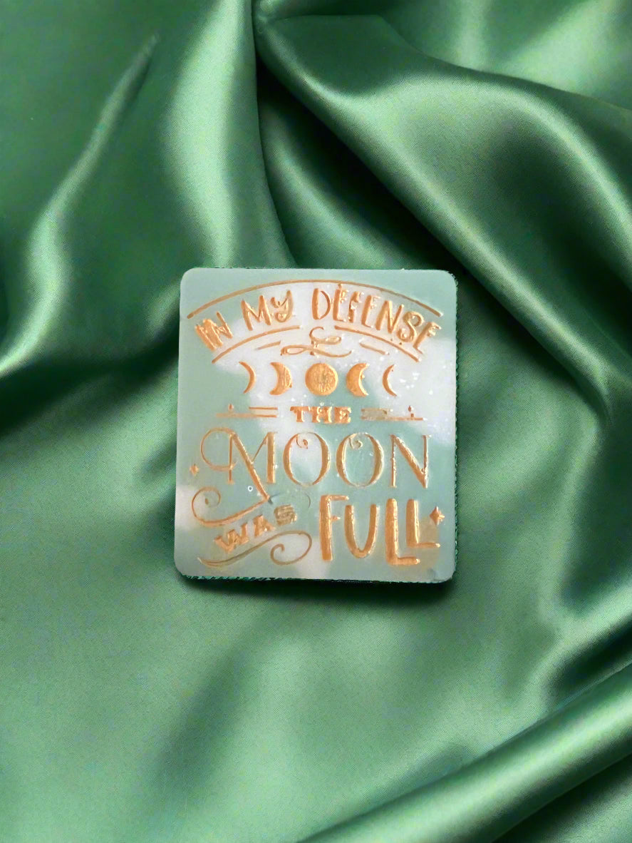 A wax melt with the text 'In my defense, the MOON WAS FULL' on it, placed on a green textured surface.