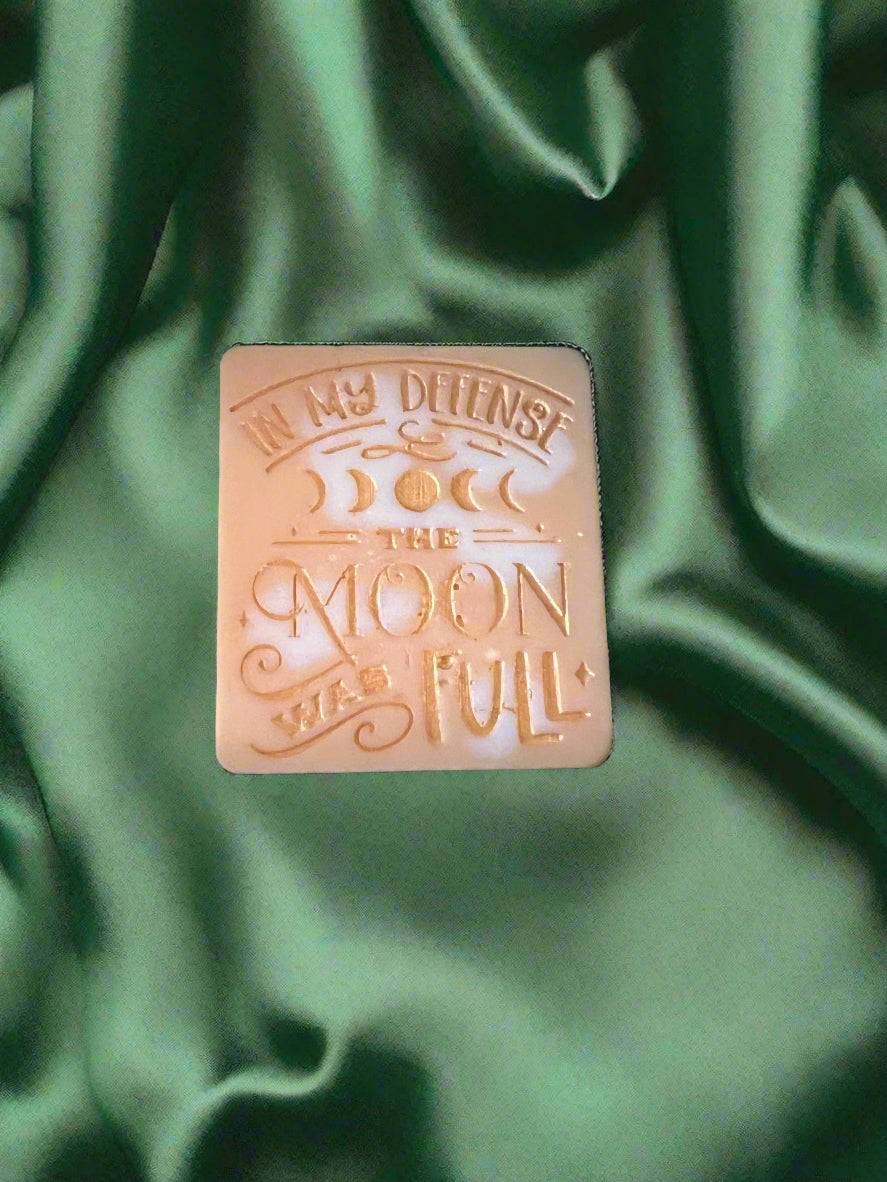 A wax melt with the text 'In my defense, the MOON WAS FULL' on it, placed on a green textured surface.
