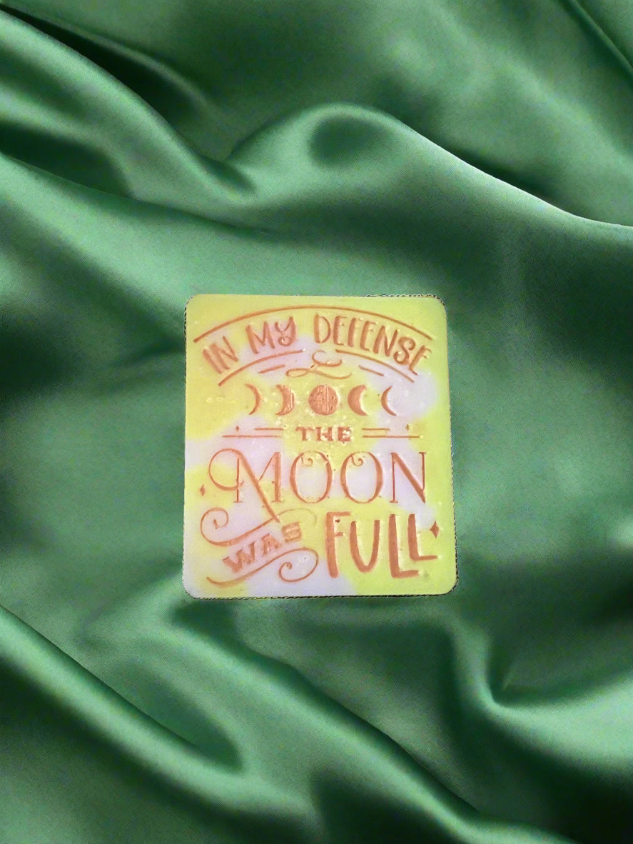 A wax melt with the text 'In my defense, the MOON WAS FULL' on it, placed on a green textured surface.
