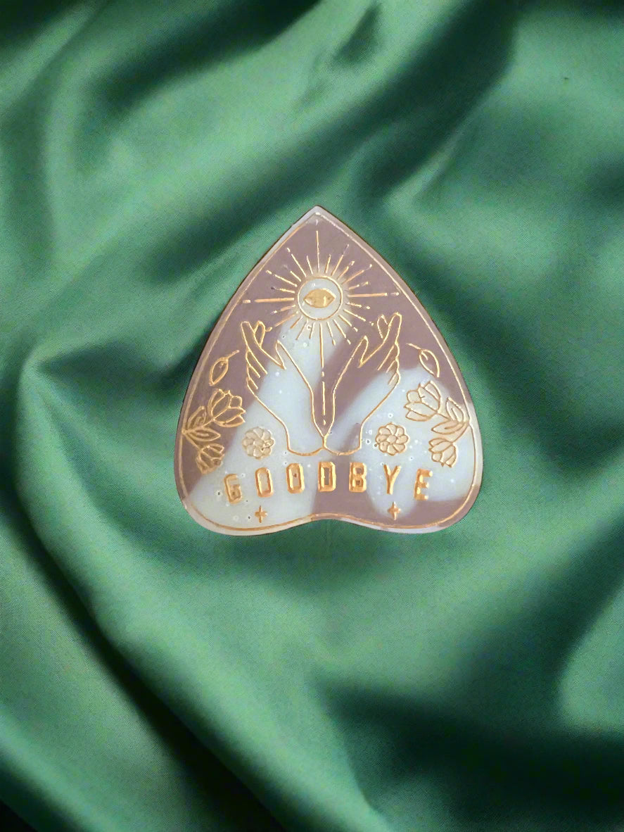 A wax melt in the shape of a planchette with a floral pattern and the word 'GOODBYE' printed on it, placed on a green textured surface.