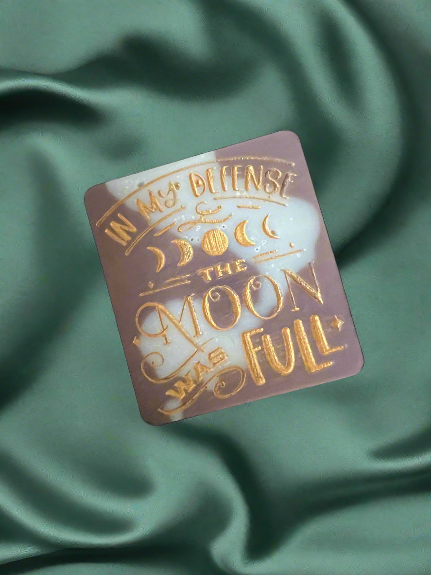 A wax melt with the text 'In my defense, the MOON WAS FULL' on it, placed on a green textured surface.