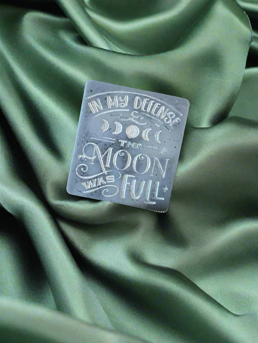 A wax melt with the text 'In my defense, the MOON WAS FULL' on it, placed on a green textured surface.