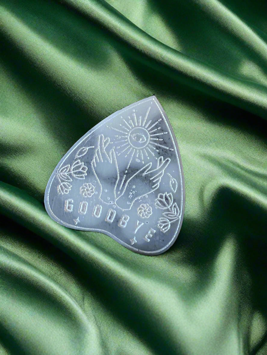 A wax melt in the shape of a planchette with a floral pattern and the word 'GOODBYE' printed on it, placed on a green textured surface.