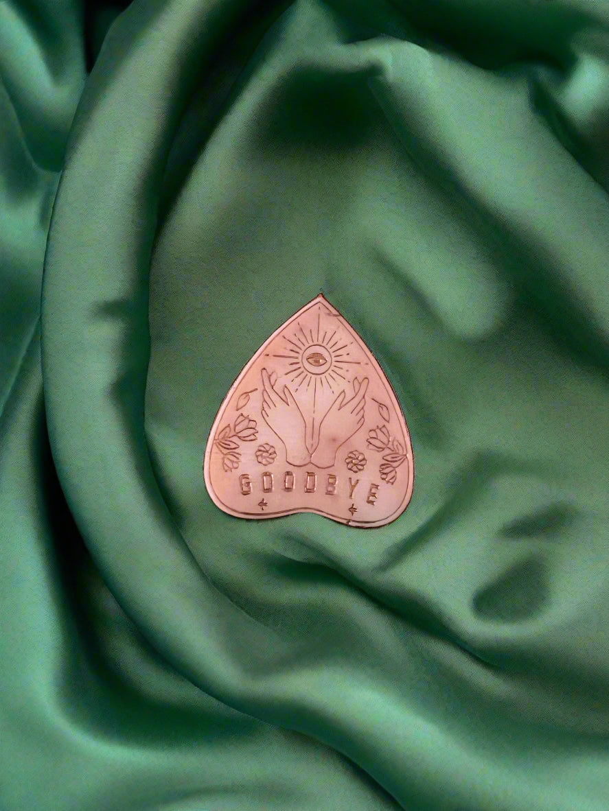 A wax melt in the shape of a planchette with a floral pattern and the word 'GOODBYE' printed on it, placed on a green textured surface.