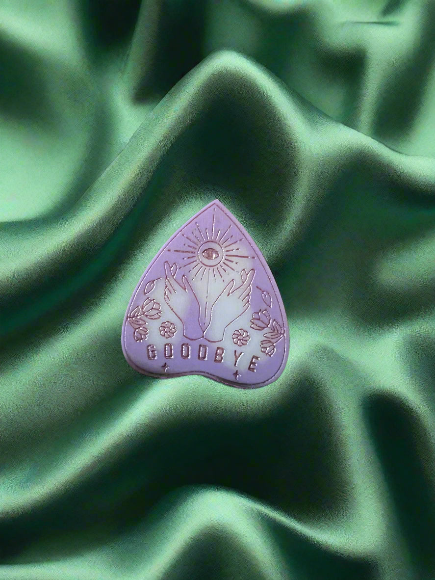 A wax melt in the shape of a planchette with a floral pattern and the word 'GOODBYE' printed on it, placed on a green textured surface.