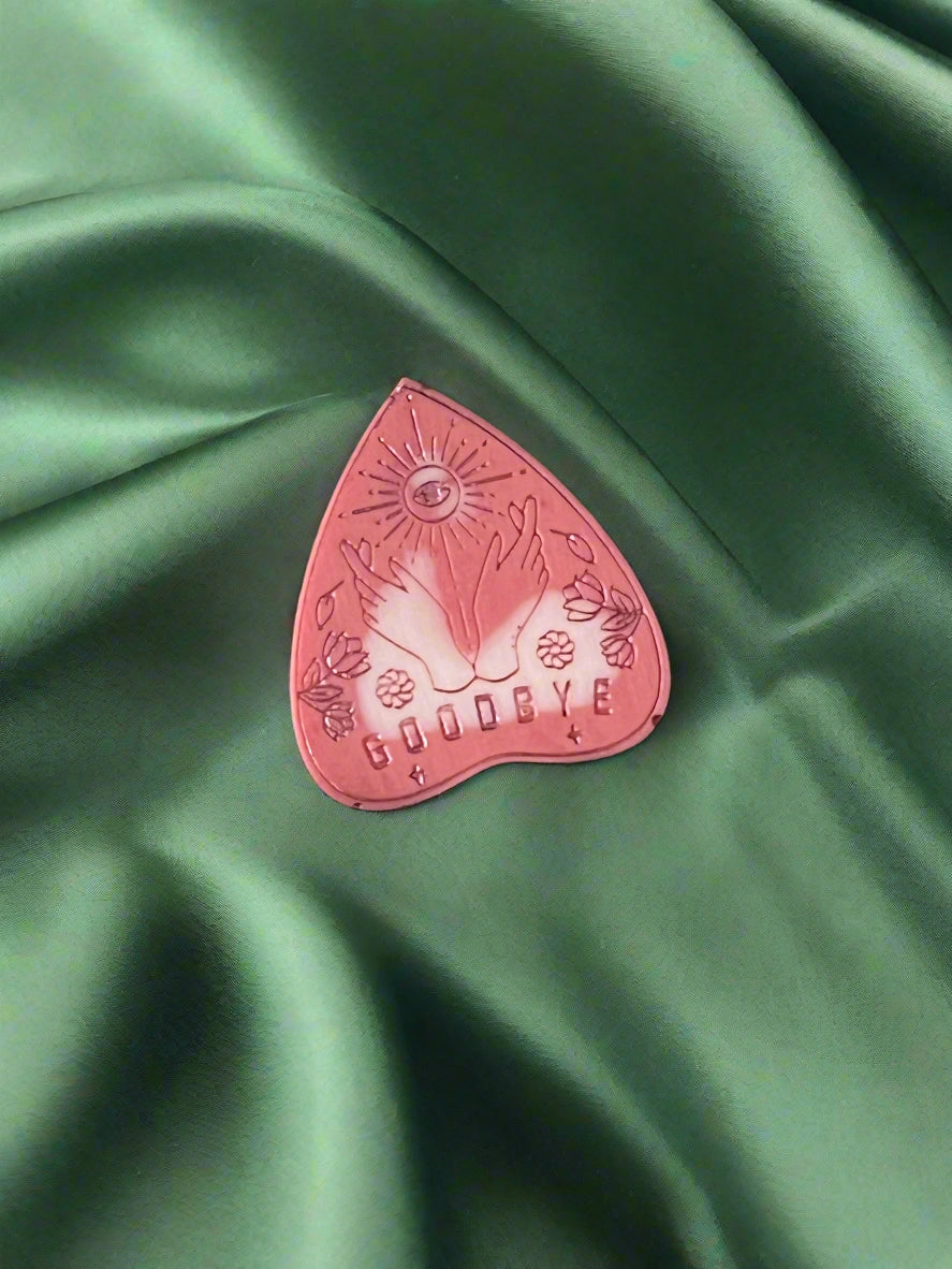 A wax melt in the shape of a planchette with a floral pattern and the word 'GOODBYE' printed on it, placed on a green textured surface.
