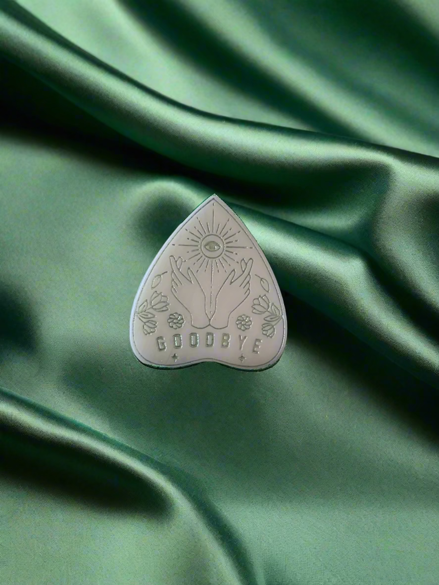 A wax melt in the shape of a planchette with a floral pattern and the word 'GOODBYE' printed on it, placed on a green textured surface.