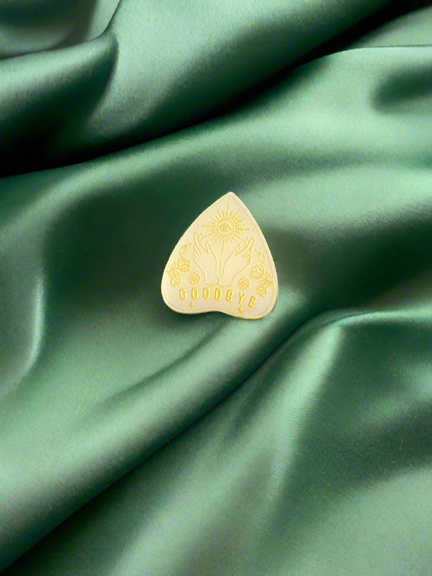 A wax melt in the shape of a planchette with a floral pattern and the word 'GOODBYE' printed on it, placed on a green textured surface.