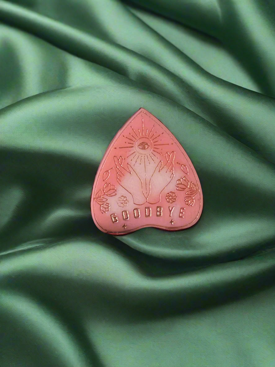 A wax melt in the shape of a planchette with a floral pattern and the word 'GOODBYE' printed on it, placed on a green textured surface.